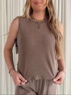 Woman wearing a brown sleeveless top and pants against a white curtain background